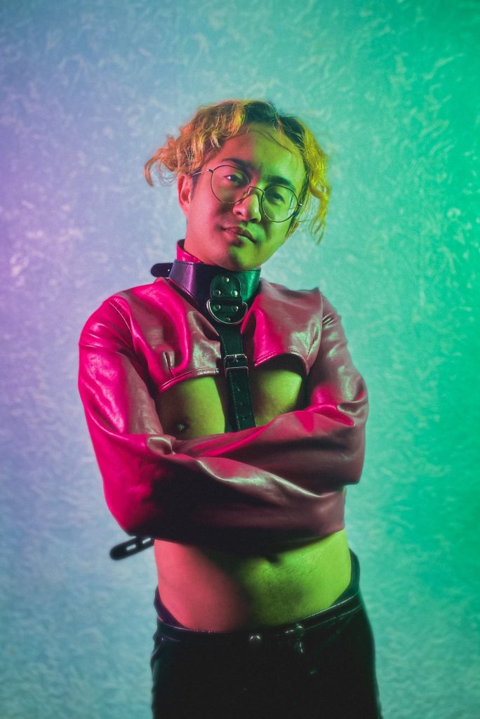 Kaya standing with a smug face while in a red straitjacket. They are lit by green and pink lights from either side for a dramatic look.